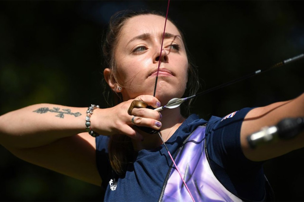 Female archer Bryony Pitman pulls the bowstring back in preparation for releasing an arrow. She is a white woman who is wearing her brunette hair back in a ponytail. She wears a navy blue short sleeve shirt, a silver bracelet and has a tattoo of an arrow on her right forearm.