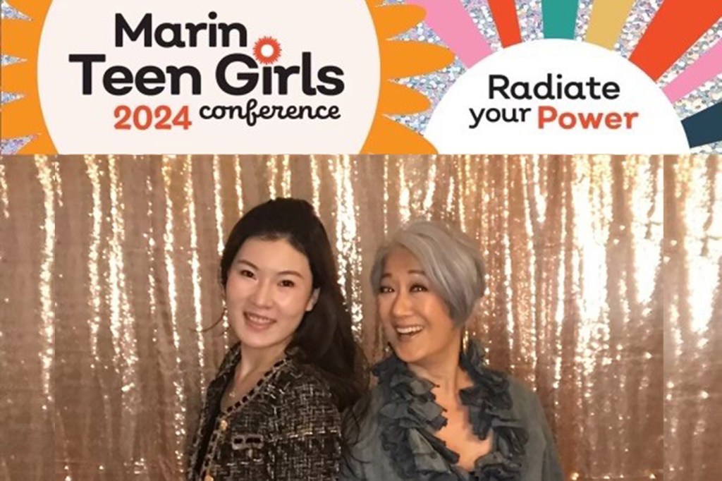 Two women are posing for a photo together. The woman on the left has long dark brown hair and brown eyes and is wearing a black and gold jacket. She is RBC Wealth Management client associate Jessica Yu. The woman on the right is financial advisor Helen Abe. Helen has short grey hair and brown eyes and is wearing a dark green ruffled top. Both women are smiling. They are standing in front of a sparkly gold backdrop.